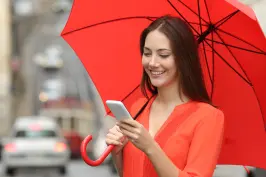 Imagem de uma moça sorrindo com um guarda-chuva laranja, roupa laranja e um celular nas mãos.