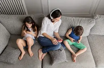 Uma mãe com um casal de filhos acessando celulares sentados em um sofá cinza.