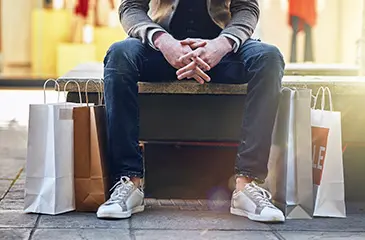 Imagem da cintura e pernas de um homem sentado em um banco cercado de sacolas de compras.